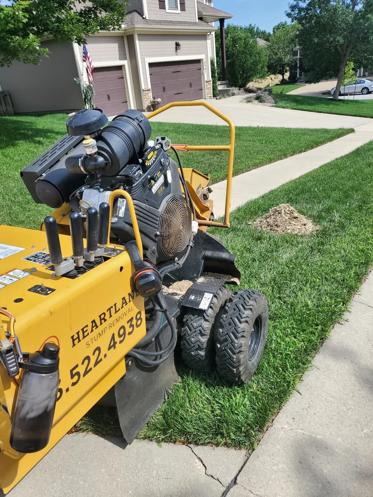 Heartland’s yellow stump grinder beside a sidewalk, with a small pile of grindings in the lawn
