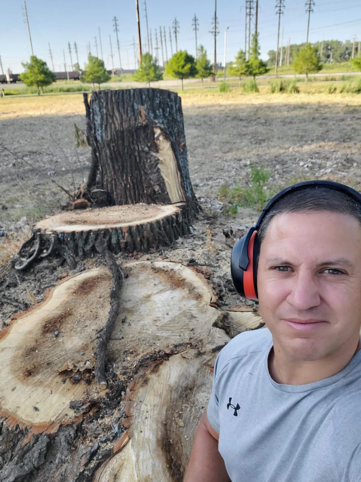 Matt wearing hearing protection beside a large remaining tree trunk and cut wood