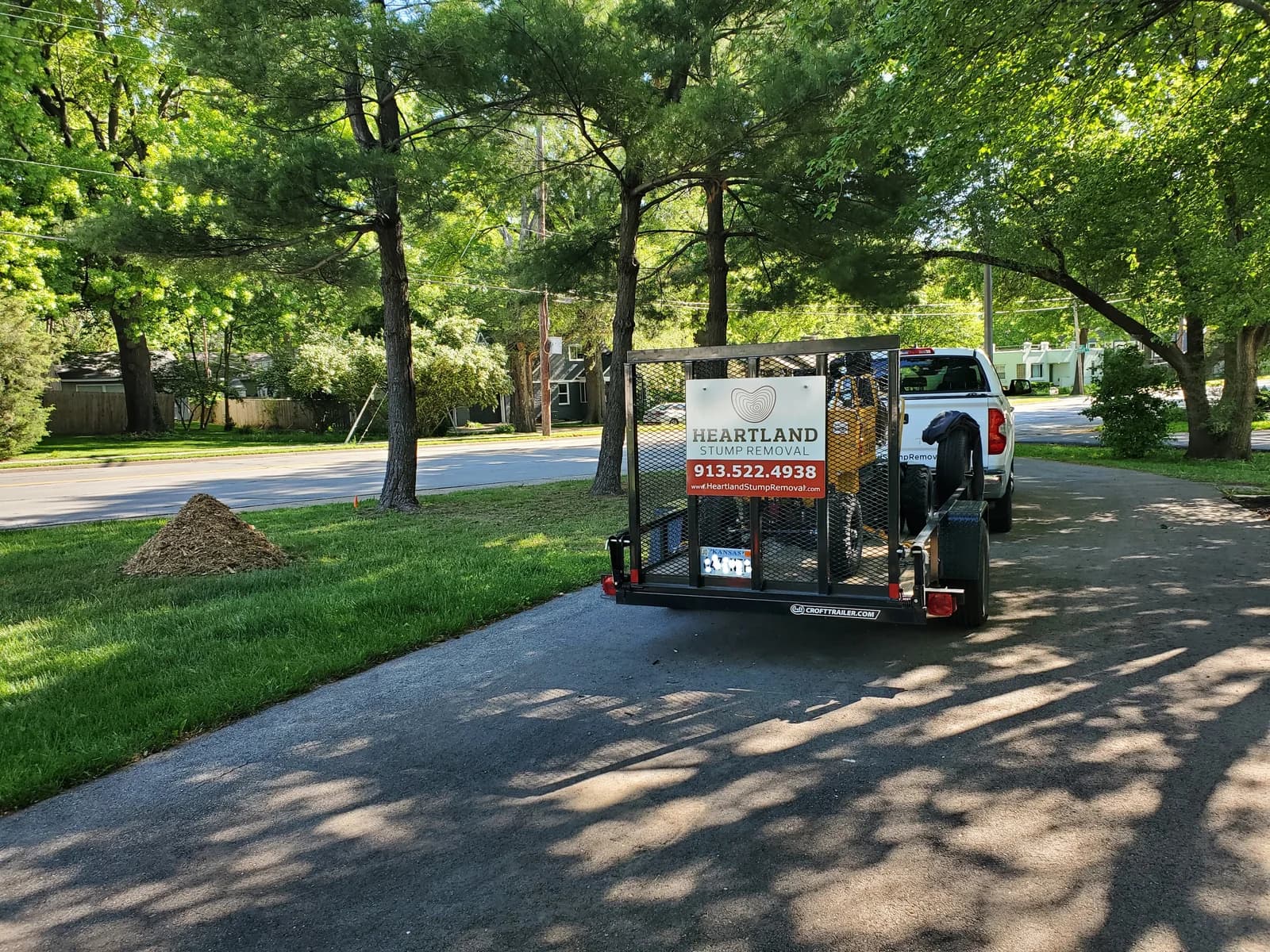 Heartland’s truck and branded trailer under shade trees beside a lawn with a pile of stump grindings