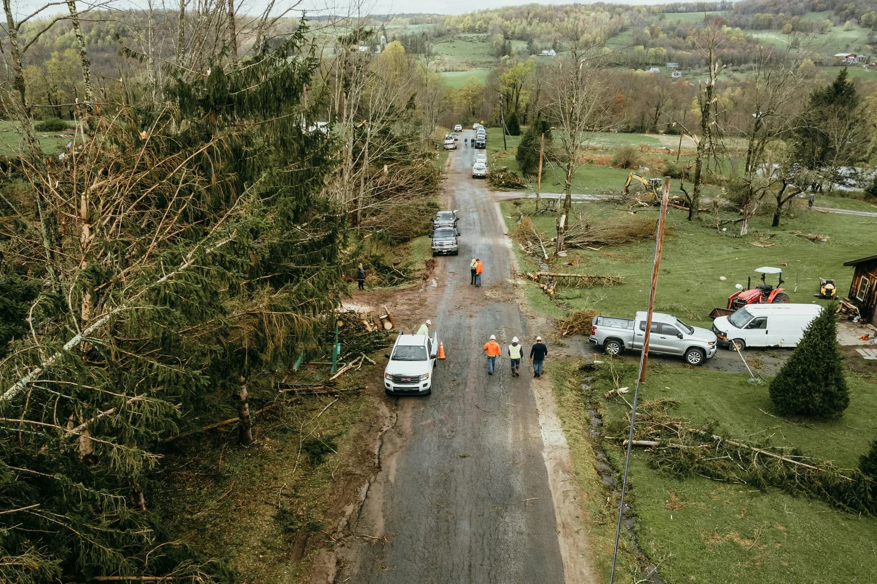 People and work vehicles along a rural road lined with broken trees and fallen branches